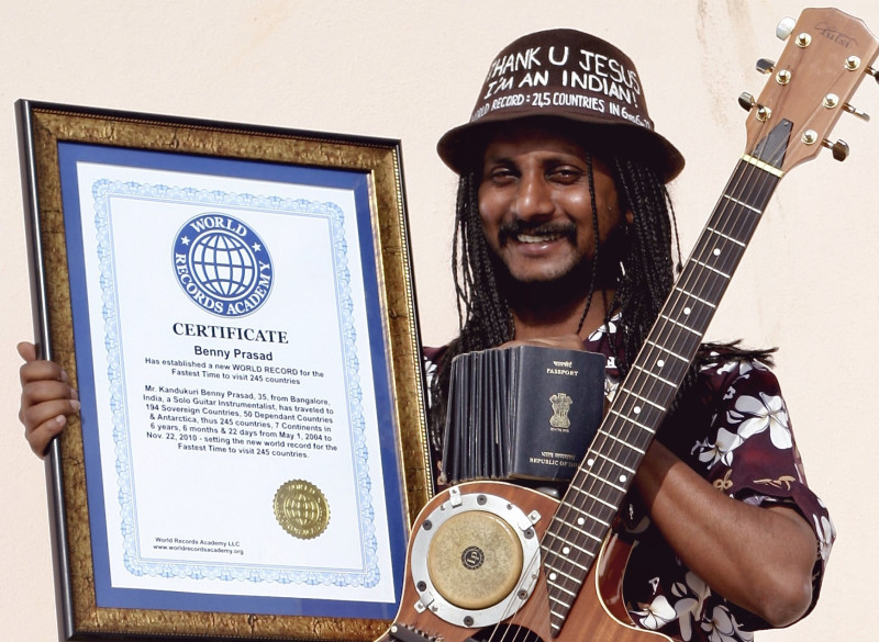 A picture of Benny Prasad, the first person from a Low Passport Idex Country, who visited every country in the world, holding a trophy, all his passports and a guitar