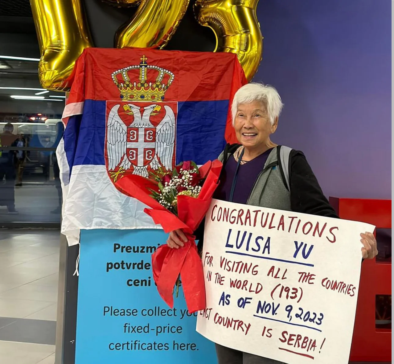 A picture of Luisa Yu, a Philipino traveller who visited every country in the world, holding her certificate upon her completion of this goal in her final country, Serbia. She is standing in front of a Serbian flag.